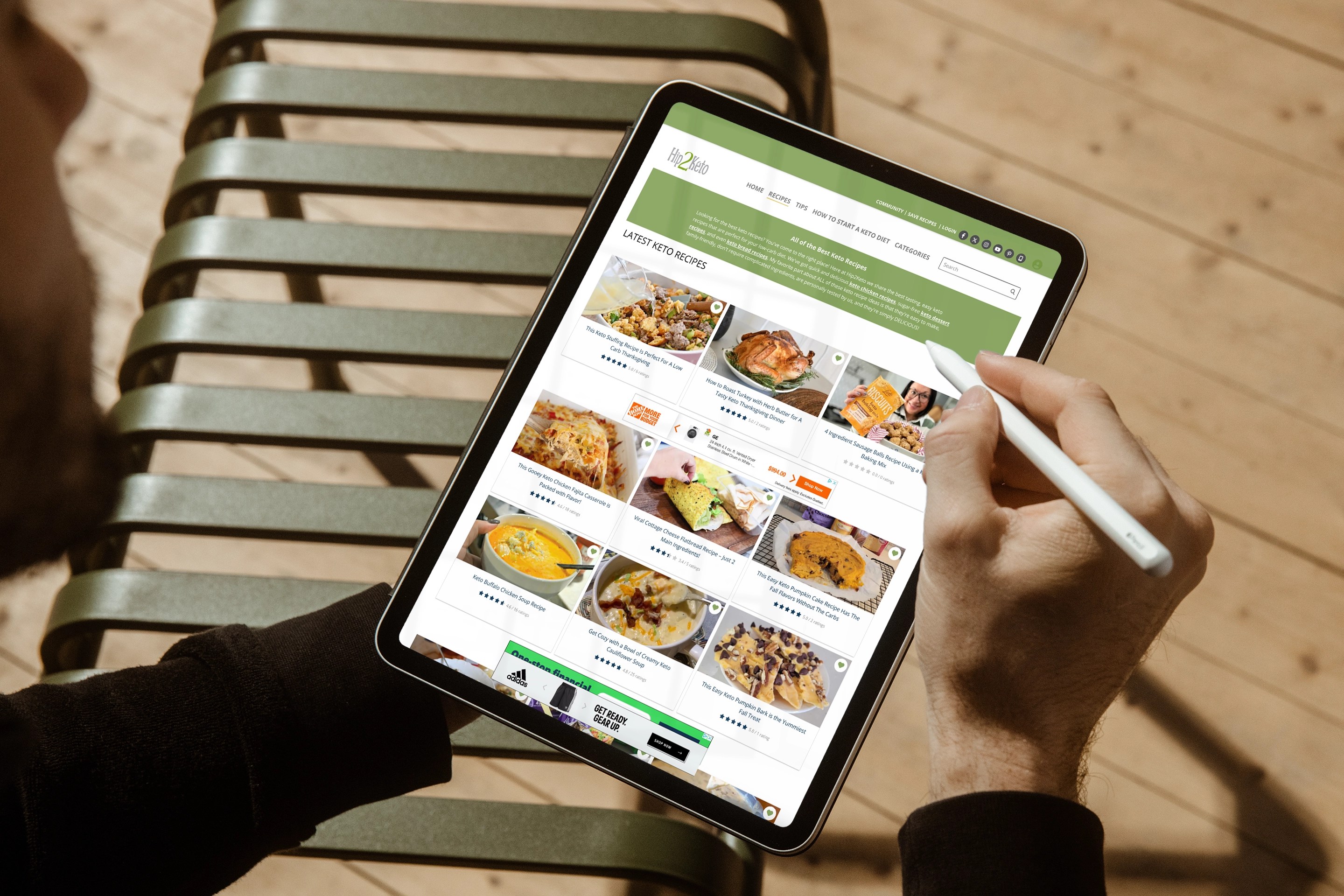 A person using a stylus on a tablet while browsing the Hip2Keto website. The screen shows a grid layout of keto-friendly recipes with thumbnails, star ratings, and titles.