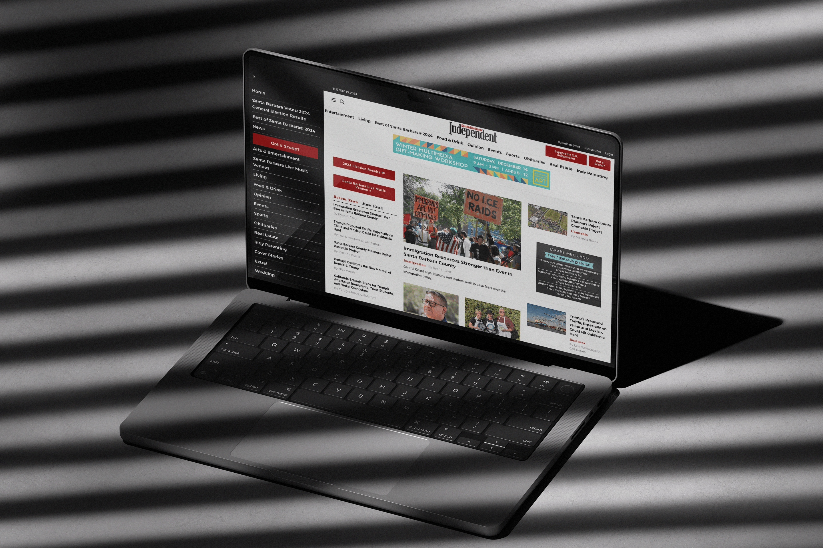 A laptop placed under striped shadows displaying the homepage of the Santa Barbara Independent website, featuring a news article about immigration resources and a sidebar with various local news categories.