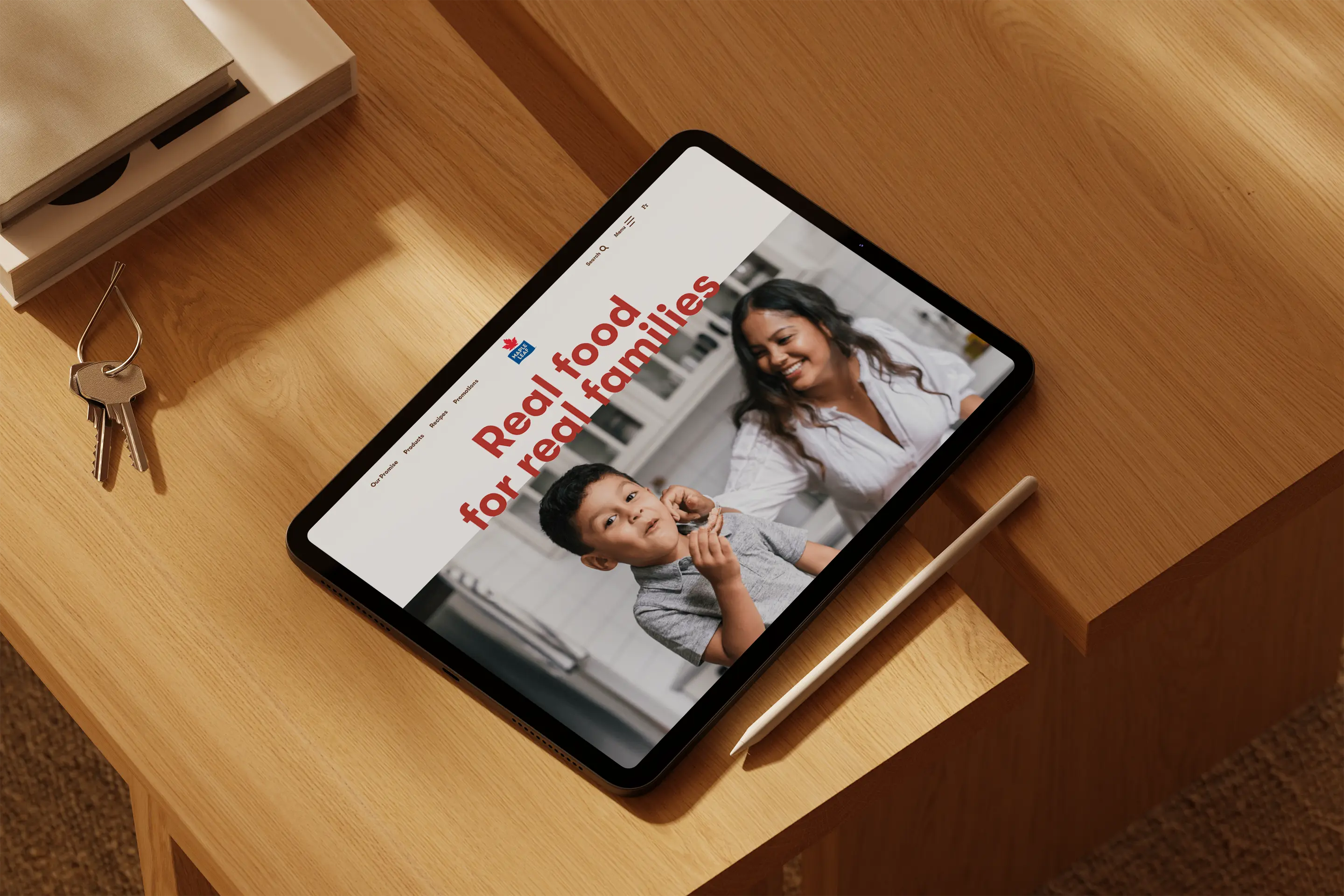 An iPad resting on a wooden table displays a webpage featuring the text "Real food for real families" alongside a smiling woman and a young boy in a kitchen setting. A stylus lies next to the tablet.