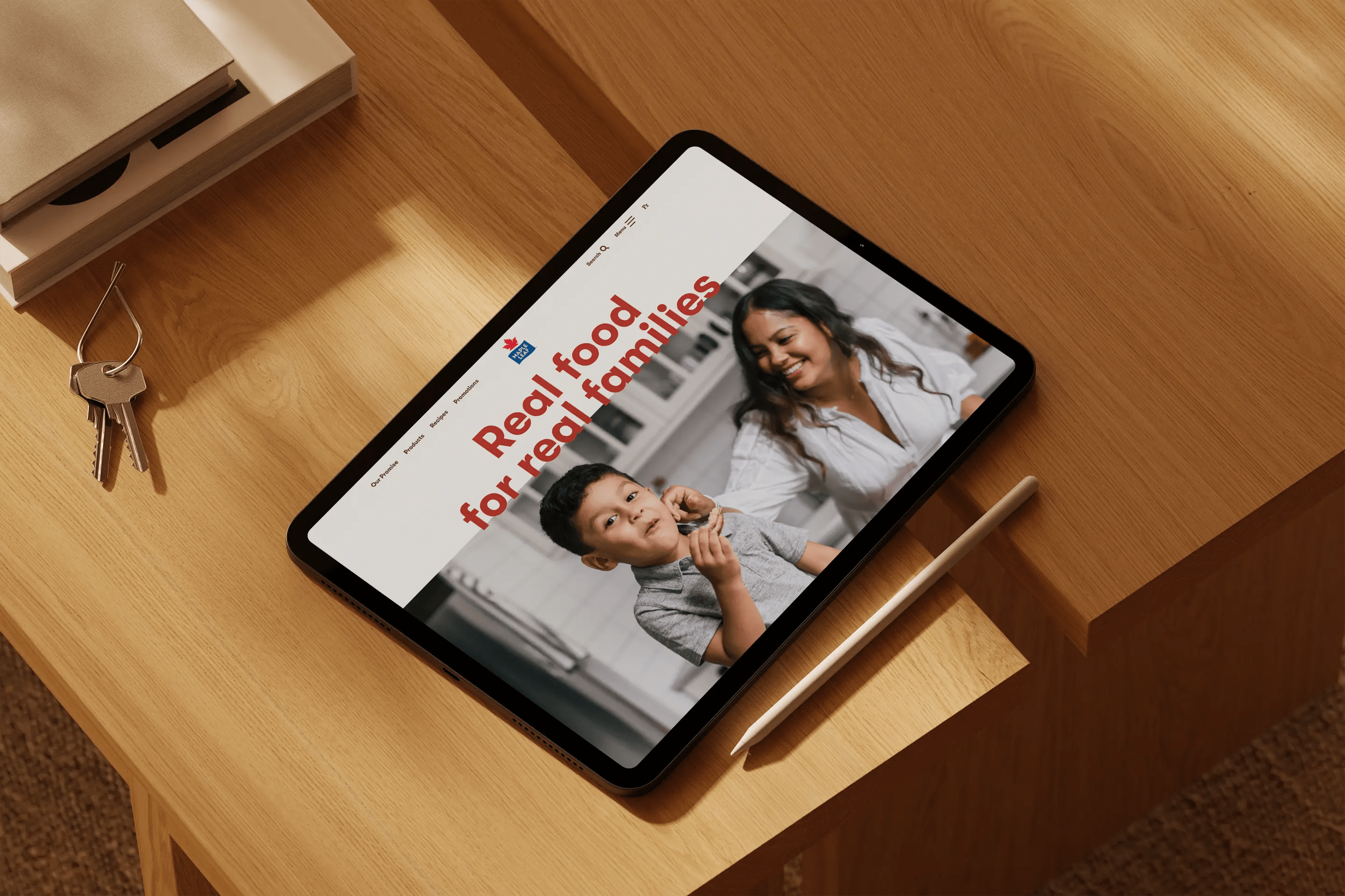 An iPad resting on a wooden table displays a webpage featuring the text "Real food for real families" alongside a smiling woman and a young boy in a kitchen setting. A stylus lies next to the tablet.