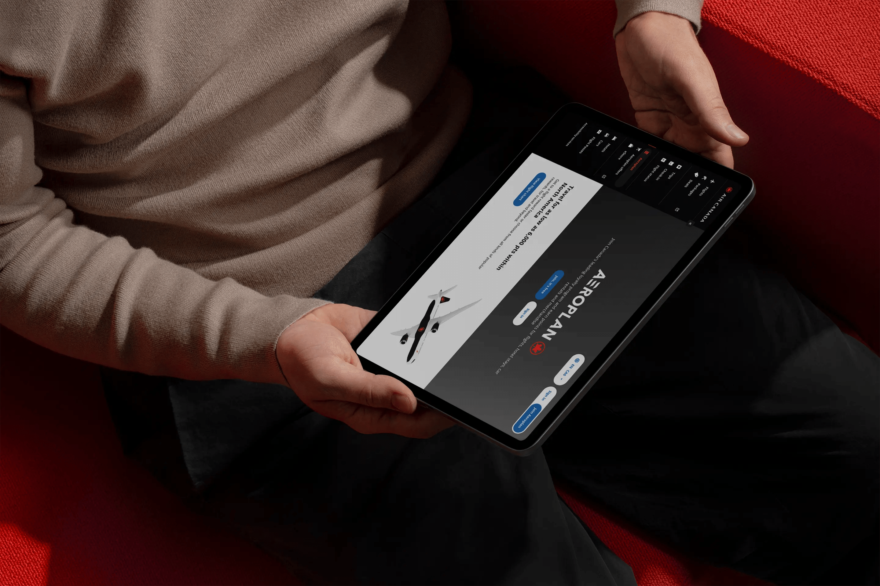 A person seated on a red couch holding a tablet horizontally, displaying the Aeroplan user dashboard. The screen shows loyalty points and travel options in a sleek dark mode UI.