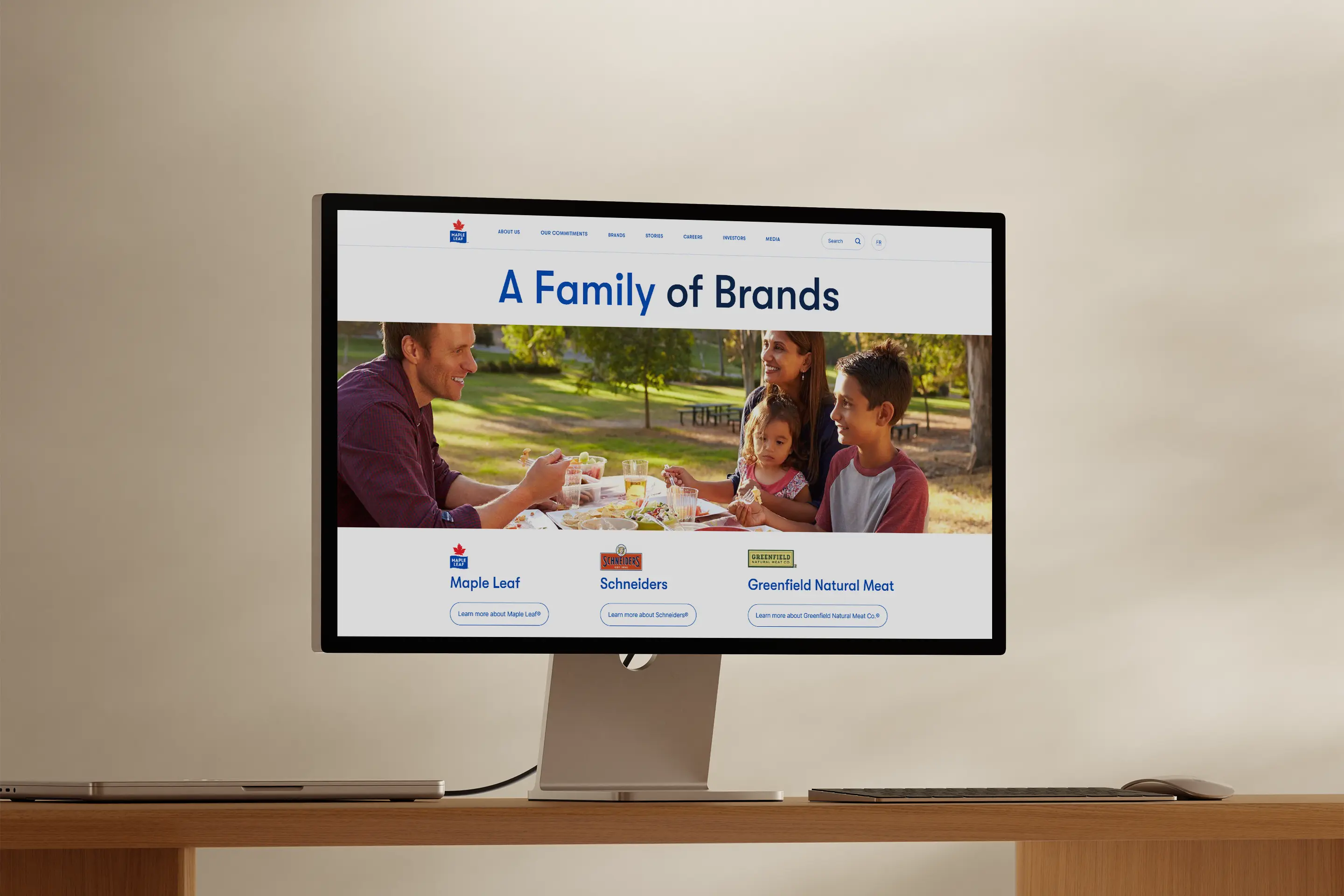 A computer monitor shows a webpage header with the text "A Family of Brands" above a family enjoying a picnic. Below, brand logos for Maple Leaf, Schneiders, and Greenfield Natural Meat are visible.