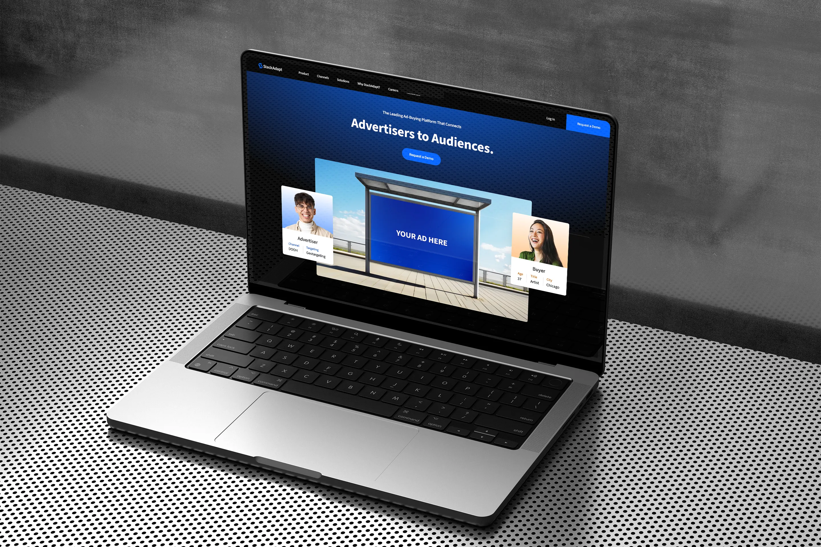 Laptop displaying the StackAdapt homepage with the tagline “Advertisers to Audiences” and a central ad mockup flanked by user profile cards, placed on a perforated metal surface.