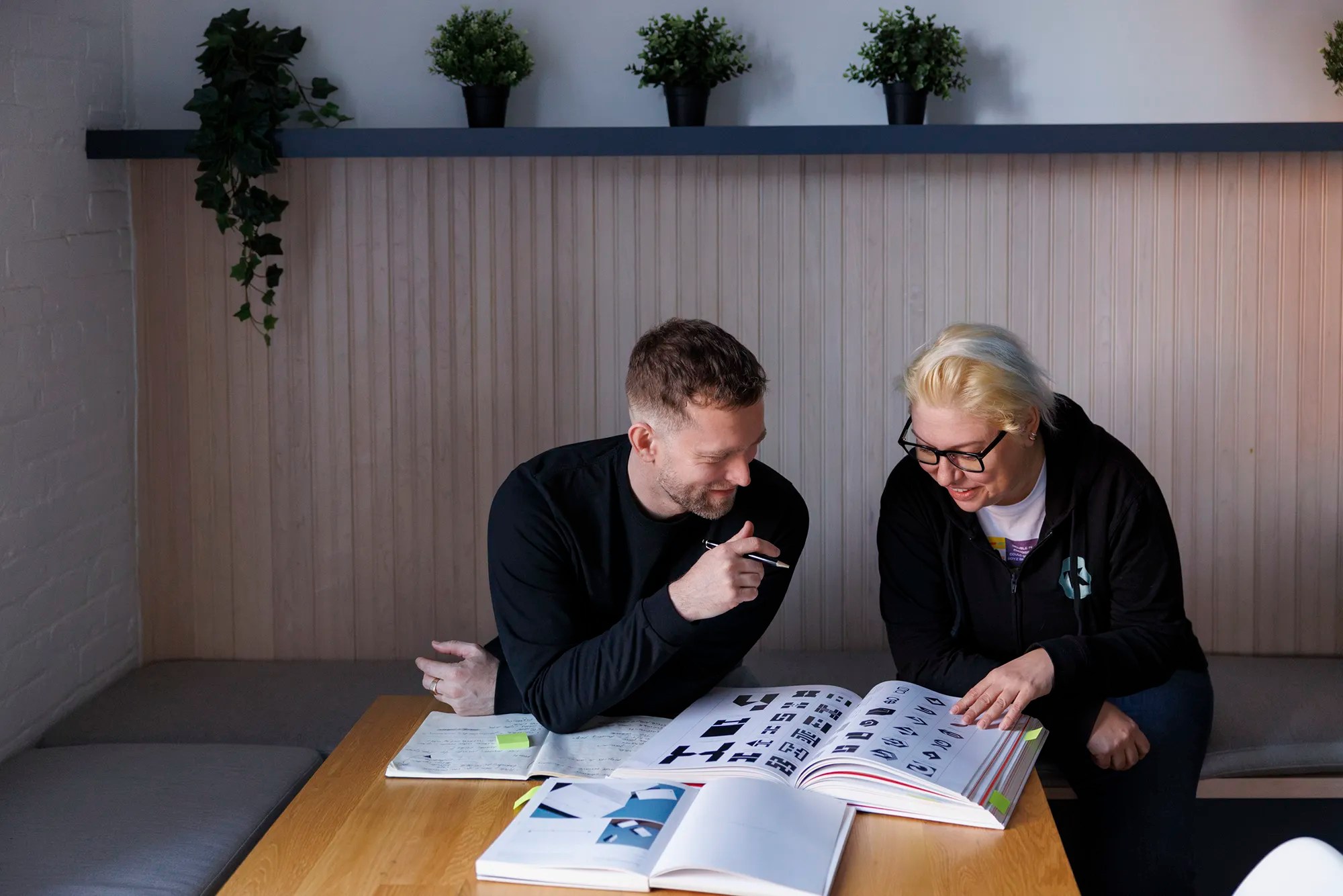 Two coworkers reviewing a book with design assets, smiling and engaged in discussion at a wooden table.