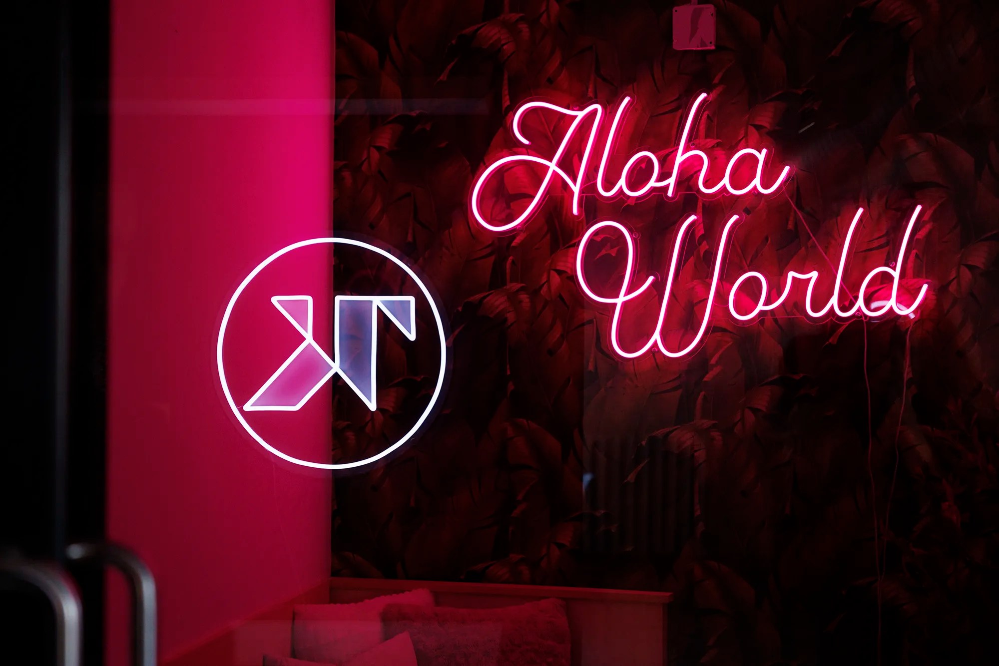 Neon sign displaying the words “Aloha World” in glowing pink script, beside a refleted Trew Knowledge logo in white, all set against a dark, leafy wall with a vibrant pink light on the left.