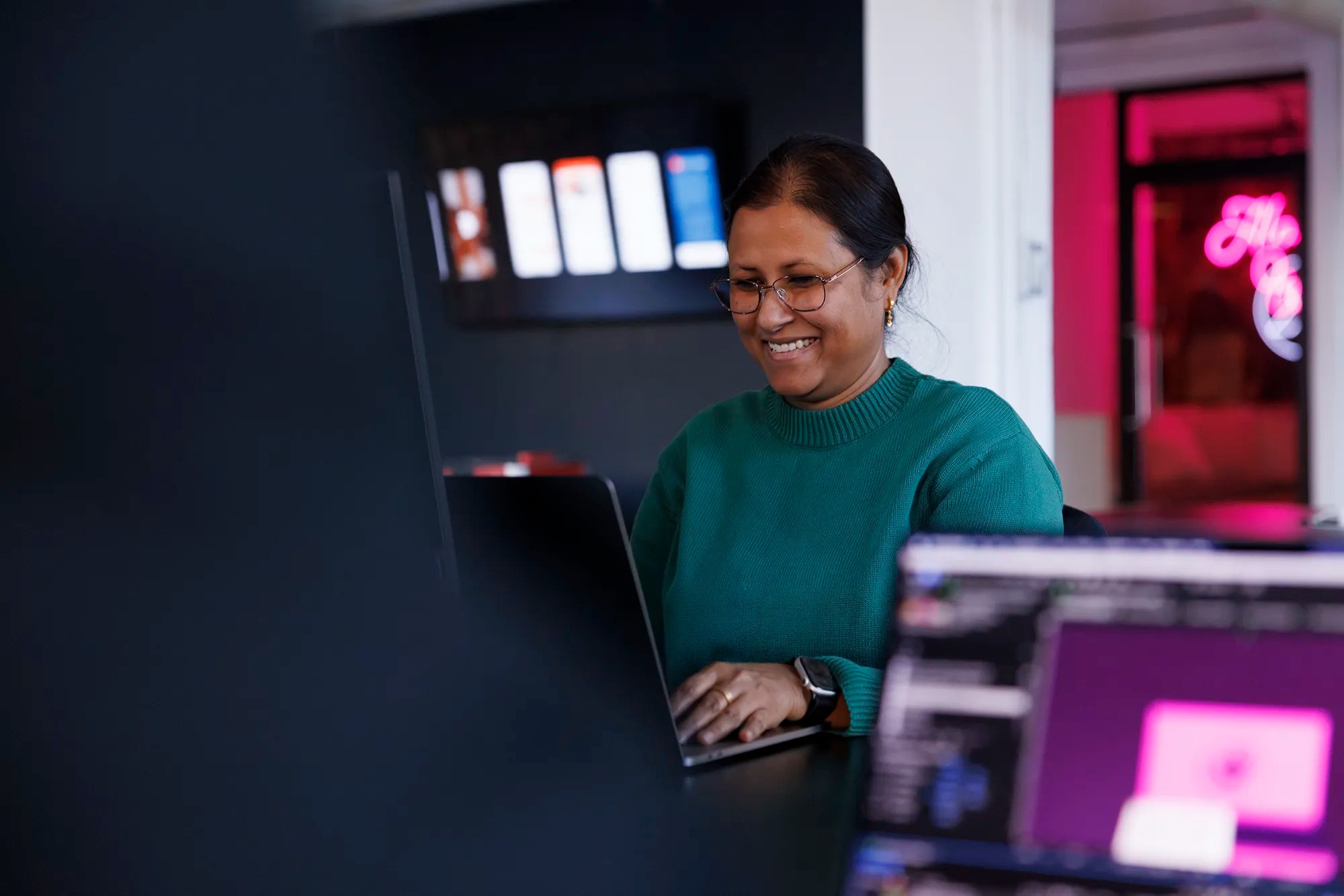 Woman in a green sweater smiling while working on a laptop, with blurred screens in the foreground and neon signage in the background.