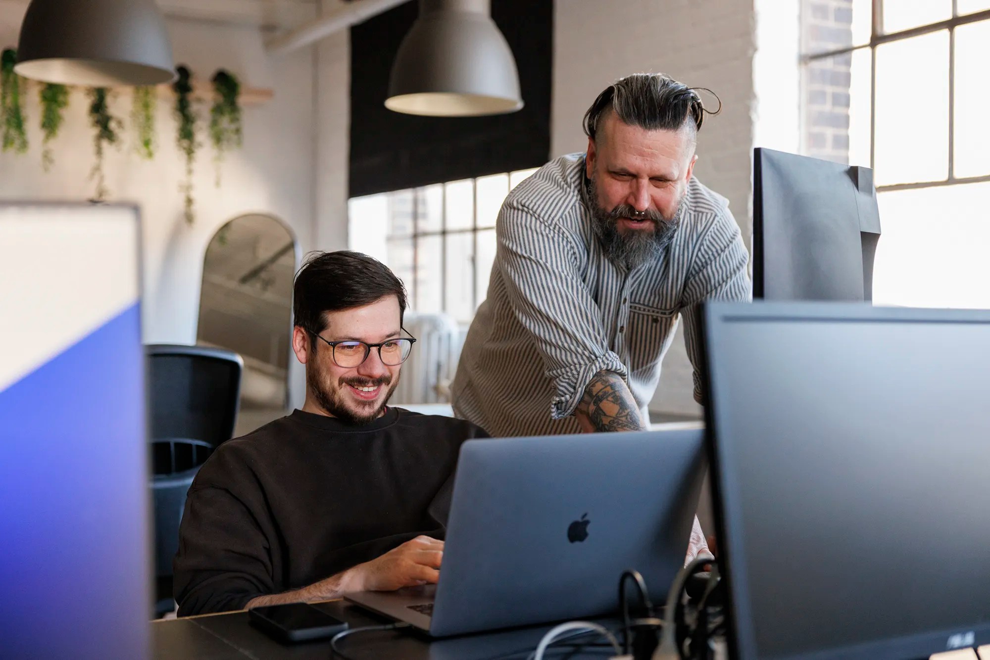 Two coworkers smiling while collaborating at a desk with a laptop in a bright, modern office space.