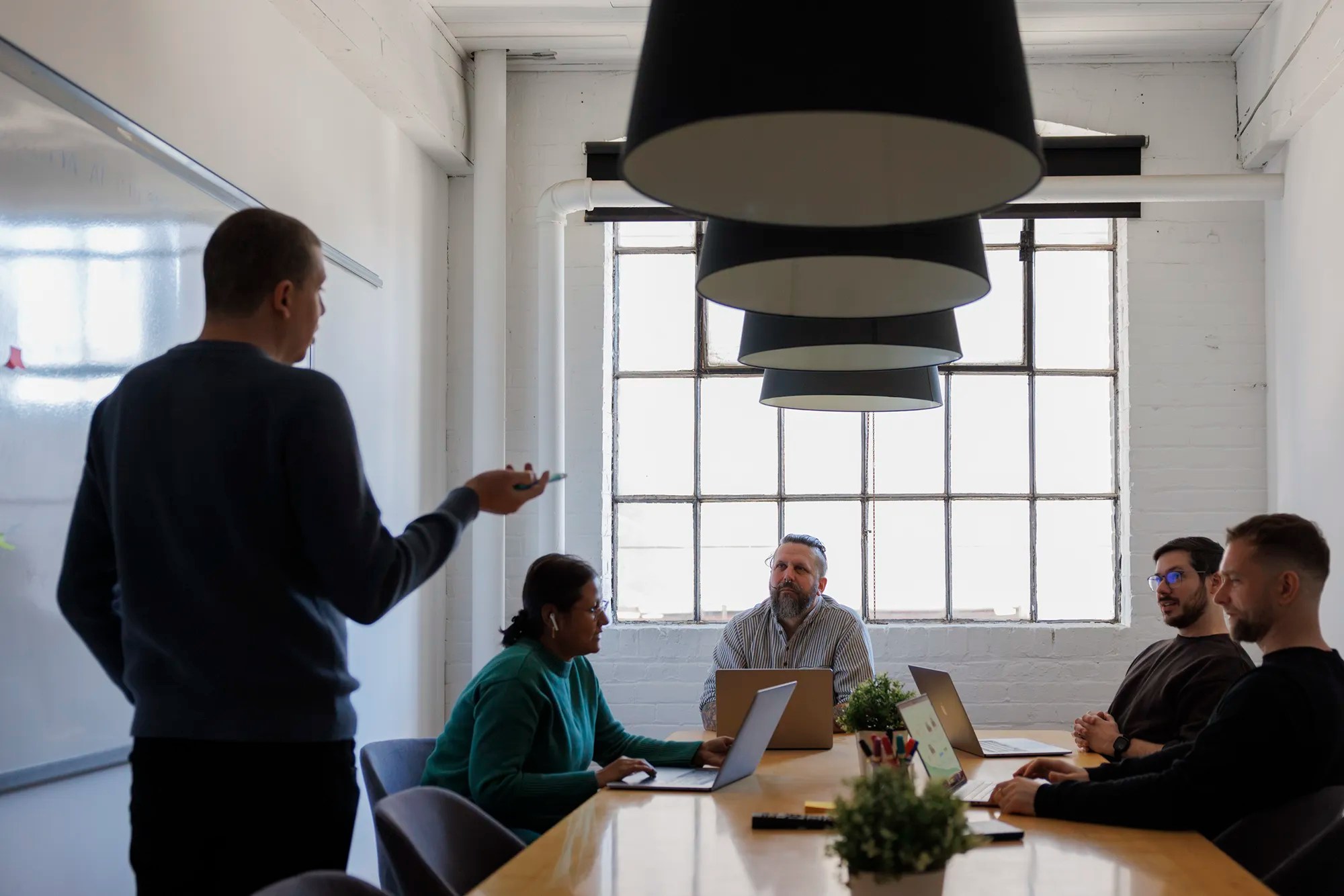 Team gathered in a conference room around a table with laptops as one person presents at the whiteboard.