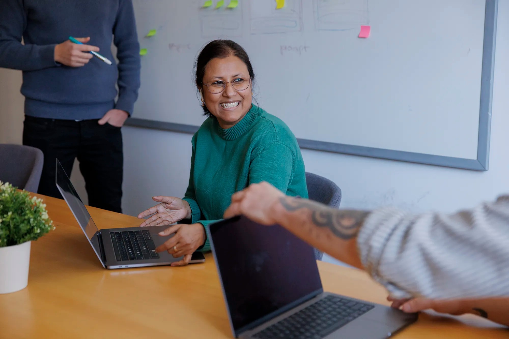 Woman smiling during a conversation in a meeting, with open laptops on the table and sticky notes on a whiteboard in the background.