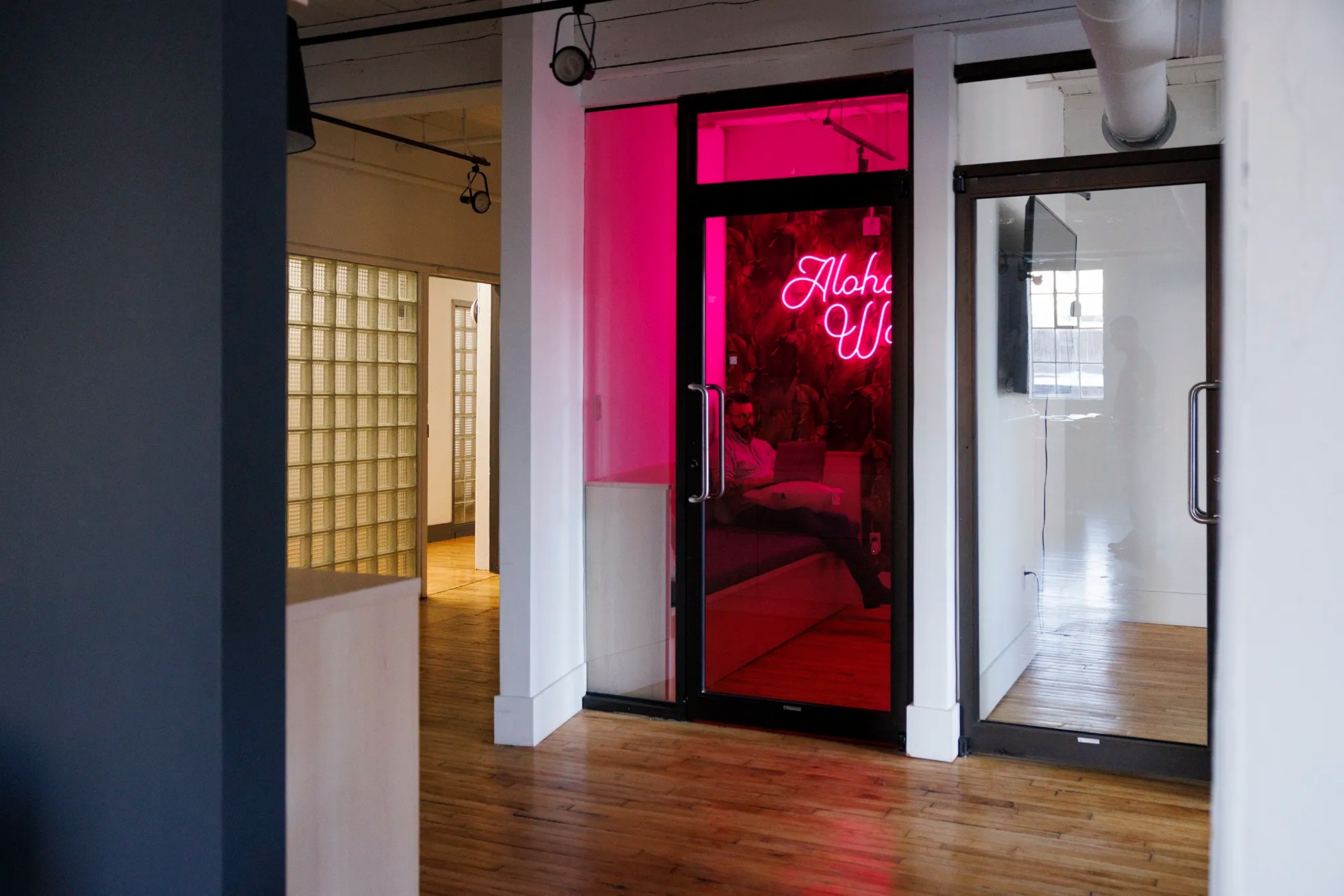 A person sitting on a couch inside a phone booth-style room with a vibrant pink neon sign reading “Aloha World” on the wall.