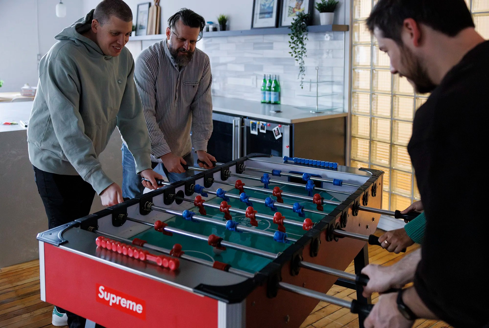 Group of coworkers playing foosball in an office kitchen area, casually enjoying a break.