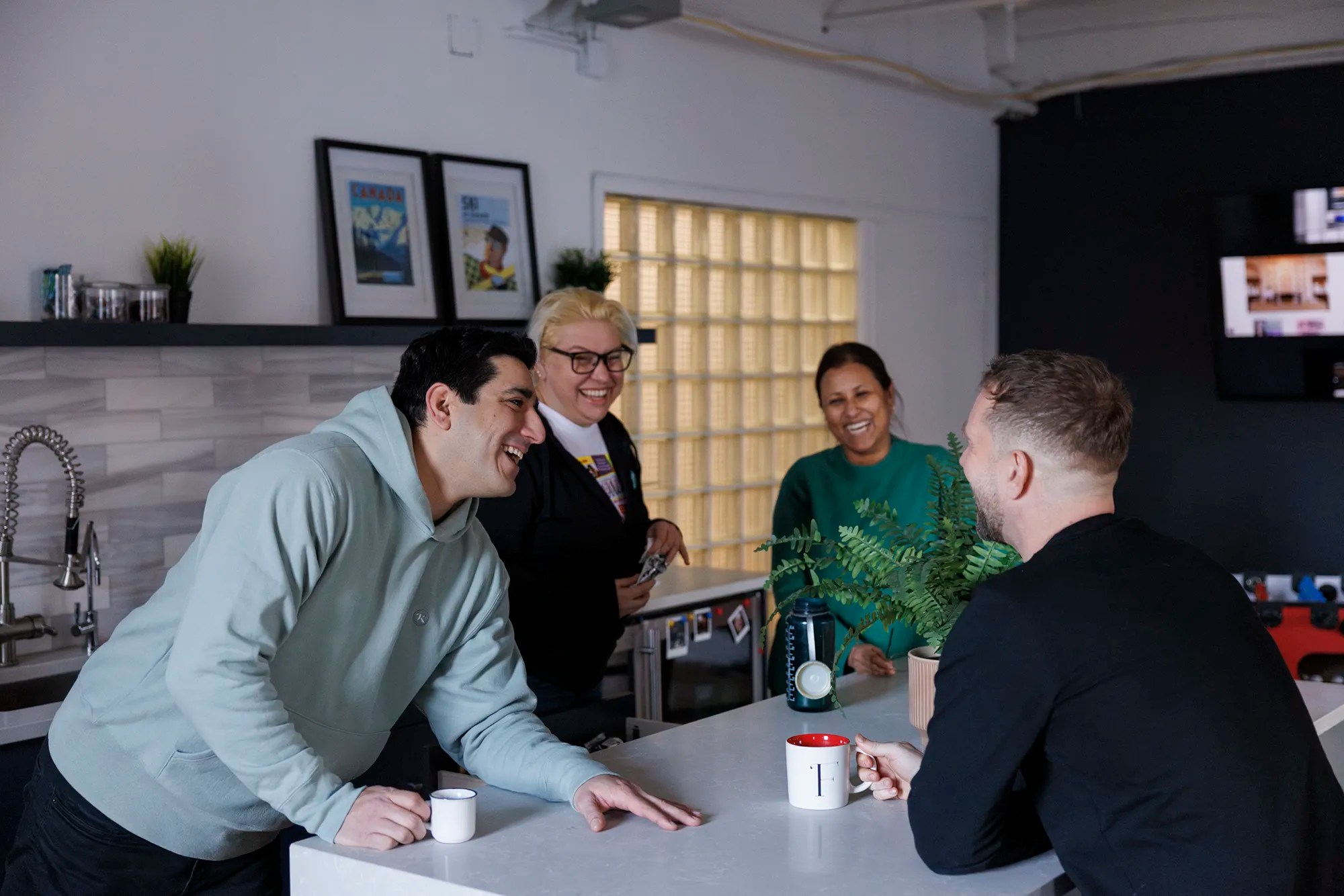 Four coworkers sharing a laugh around a white kitchen counter in a casual office setting, with mugs and plants on the counter.