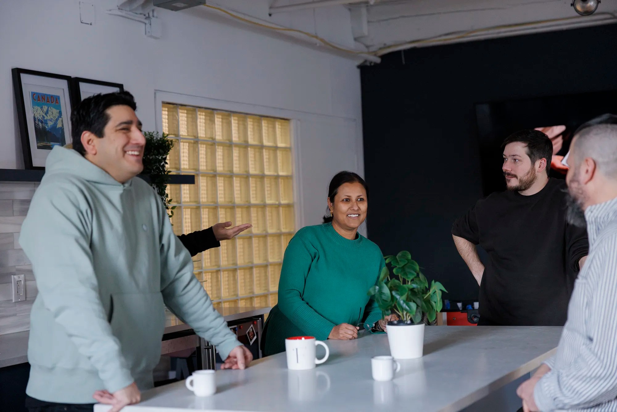 Team members chatting and laughing around a kitchen island in the office, with mugs and a potted plant on the counter.